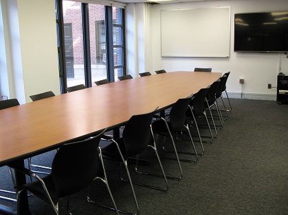 shows a small seminar room with a long oval table and chairs, and built-in AV equipment.