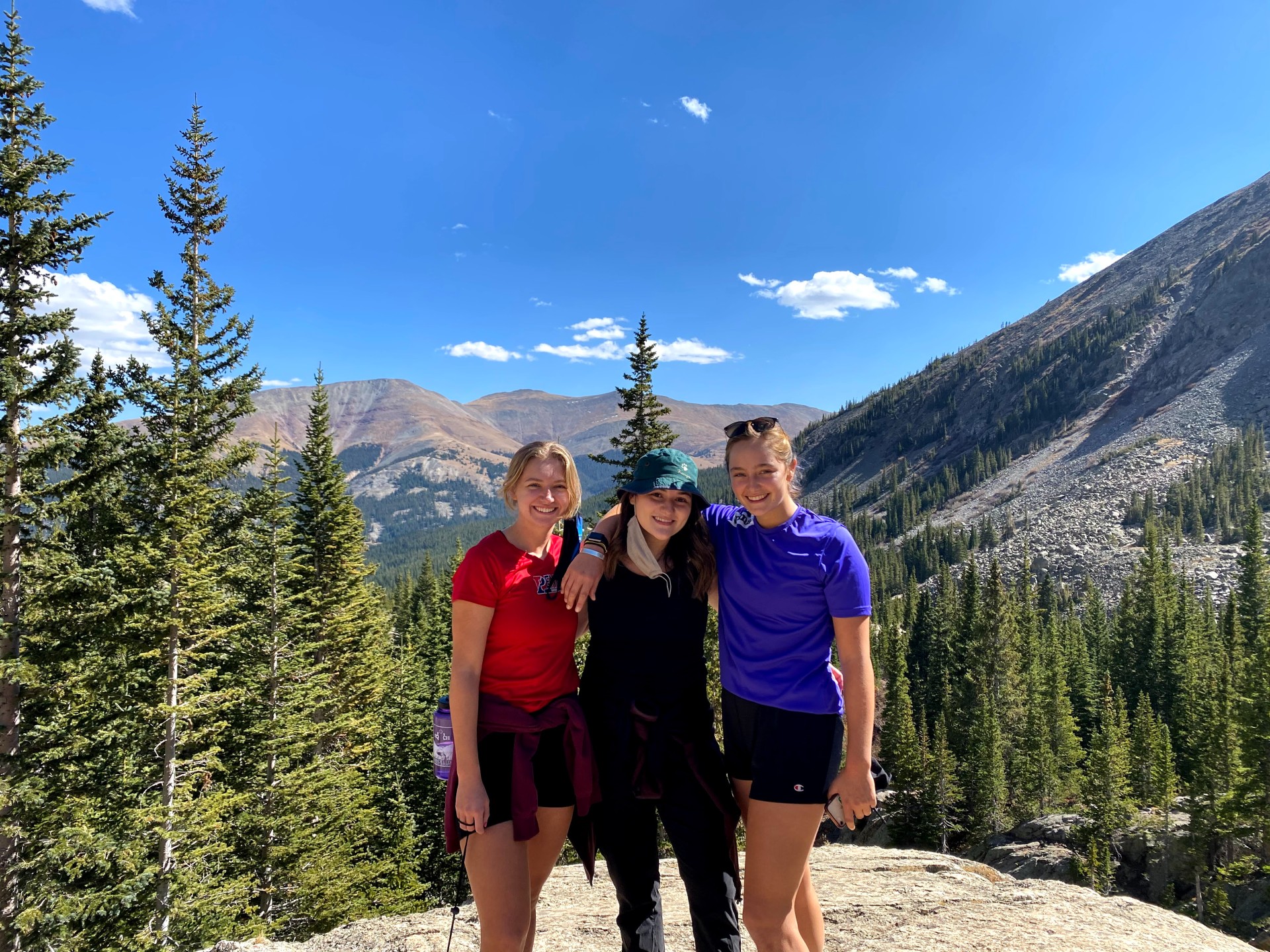 Three students standing on mountain with outlook of evergreens behind