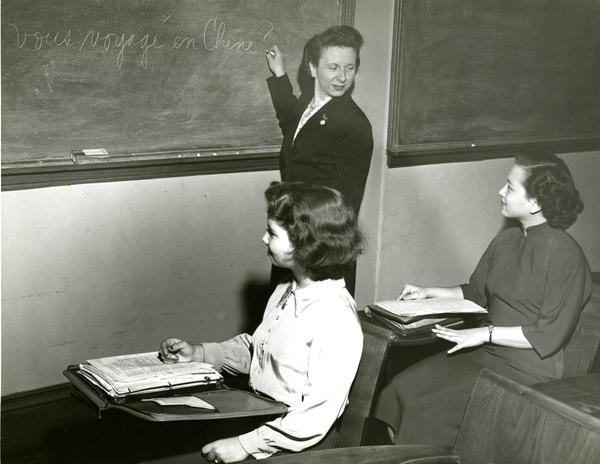 A female teacher writes on a chalkboard in front of two female students