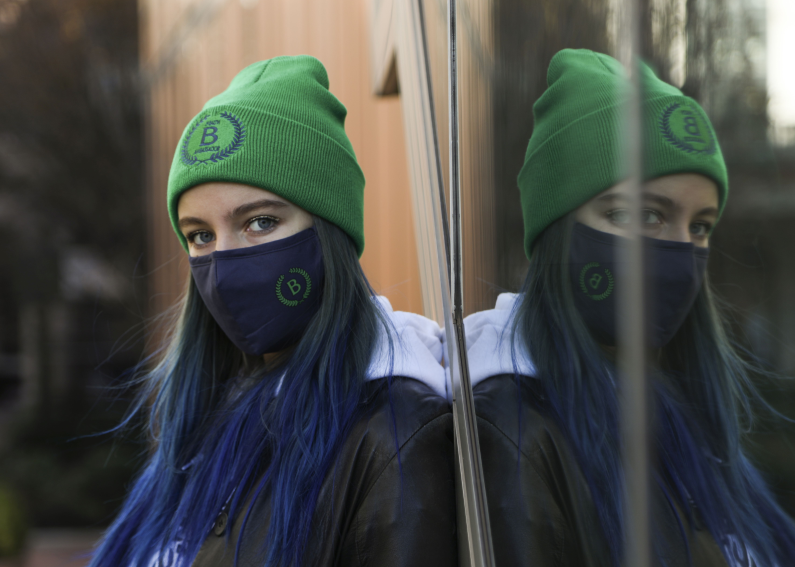 Student Audren Barbour leaning against one of the glass windows of the Diana Center with a green health ambassador beanie on,