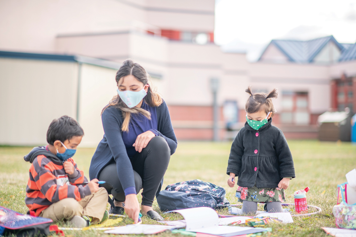 A parent colors outside with two small children, all wearing masks