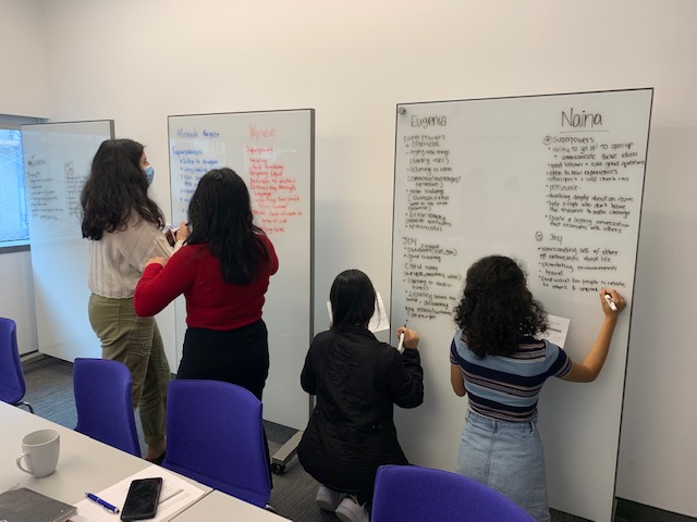Four students stand side-by-side writing on a white board and conferring with each other