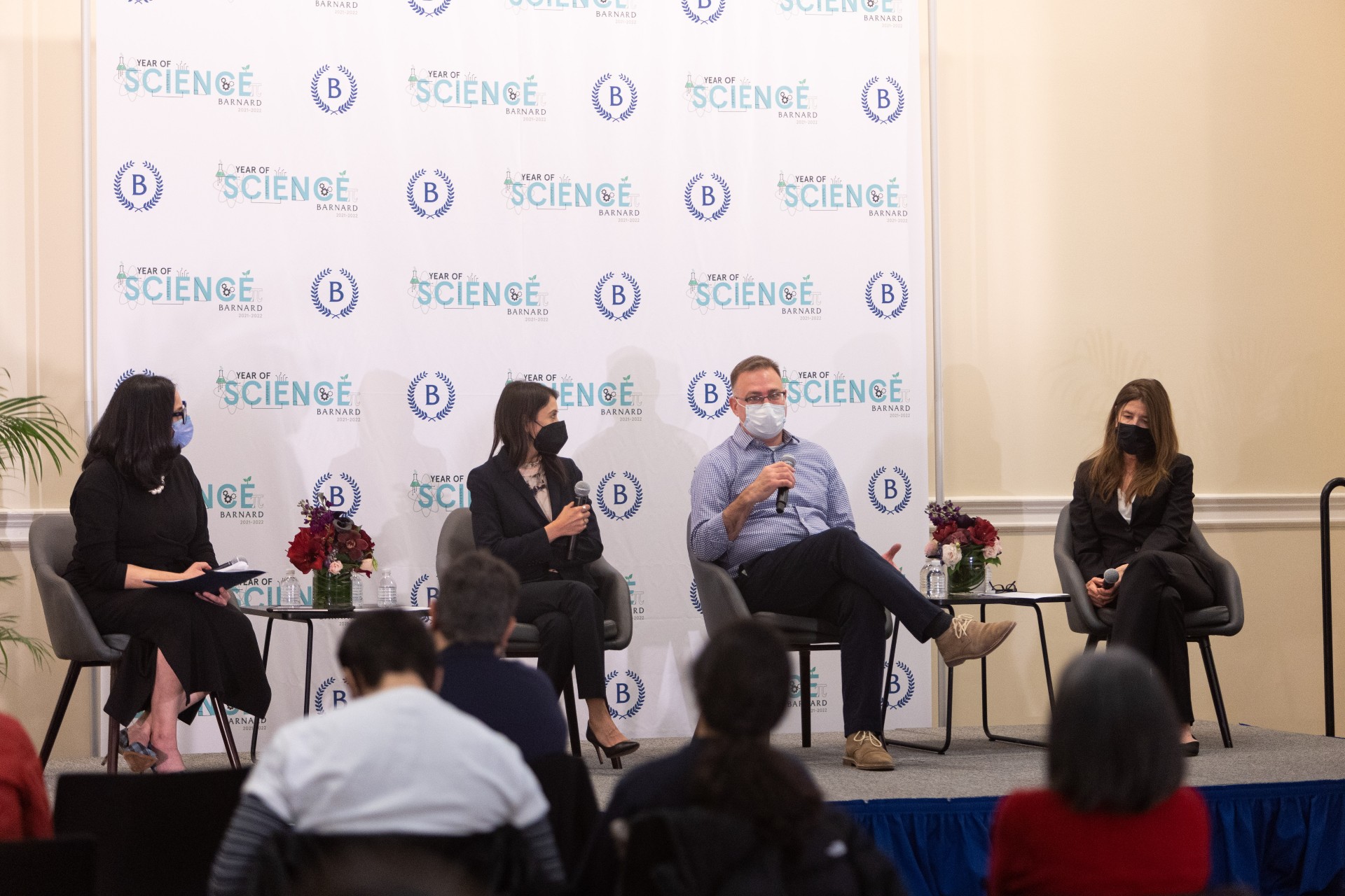 A wide shot of the 3 masked panelists and the moderator having a conversation while seated on stage in front of a Year of Science backdrop