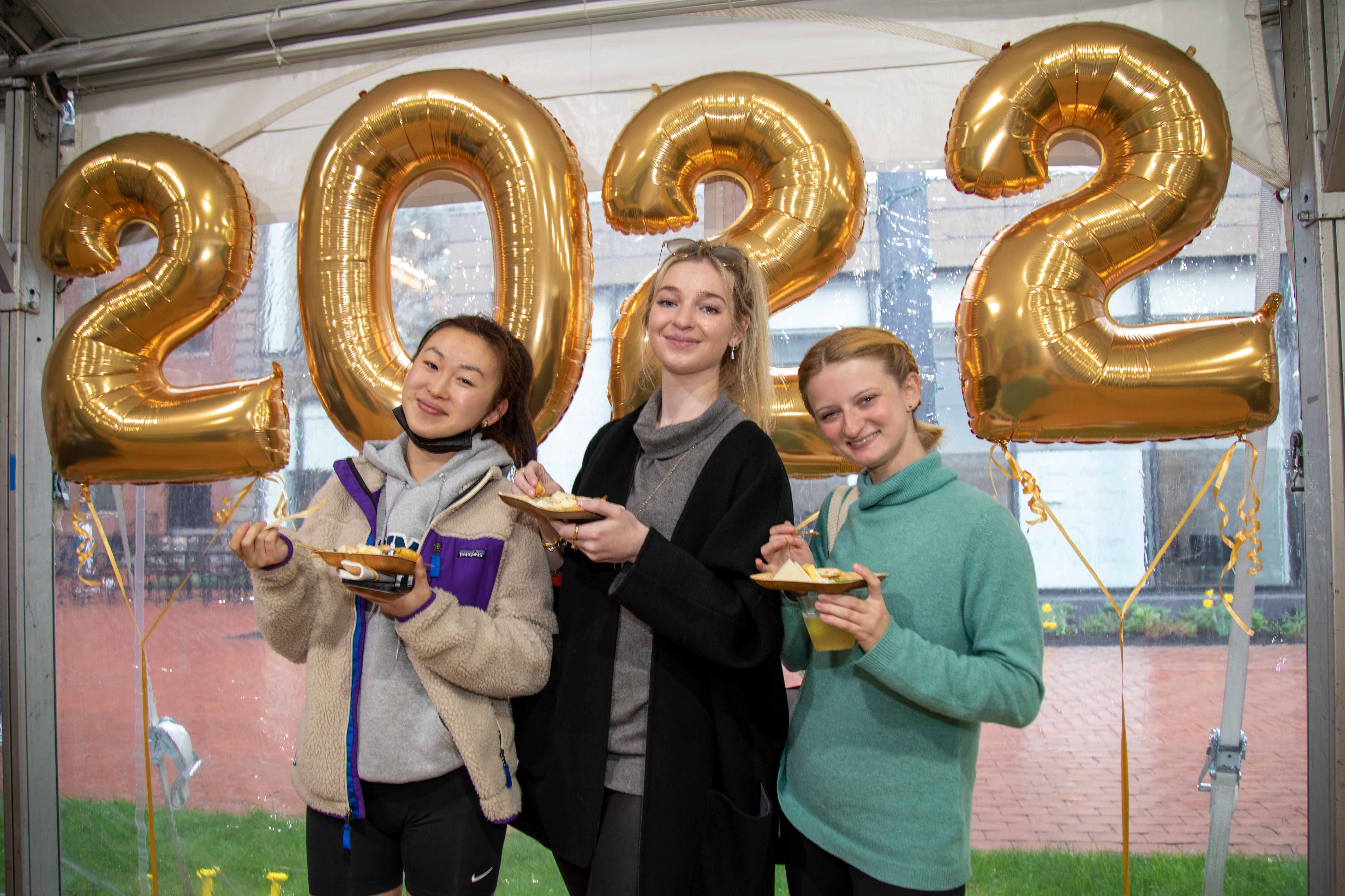 Three students pose with food in hand and 2022 balloons behind them at the senior reception.