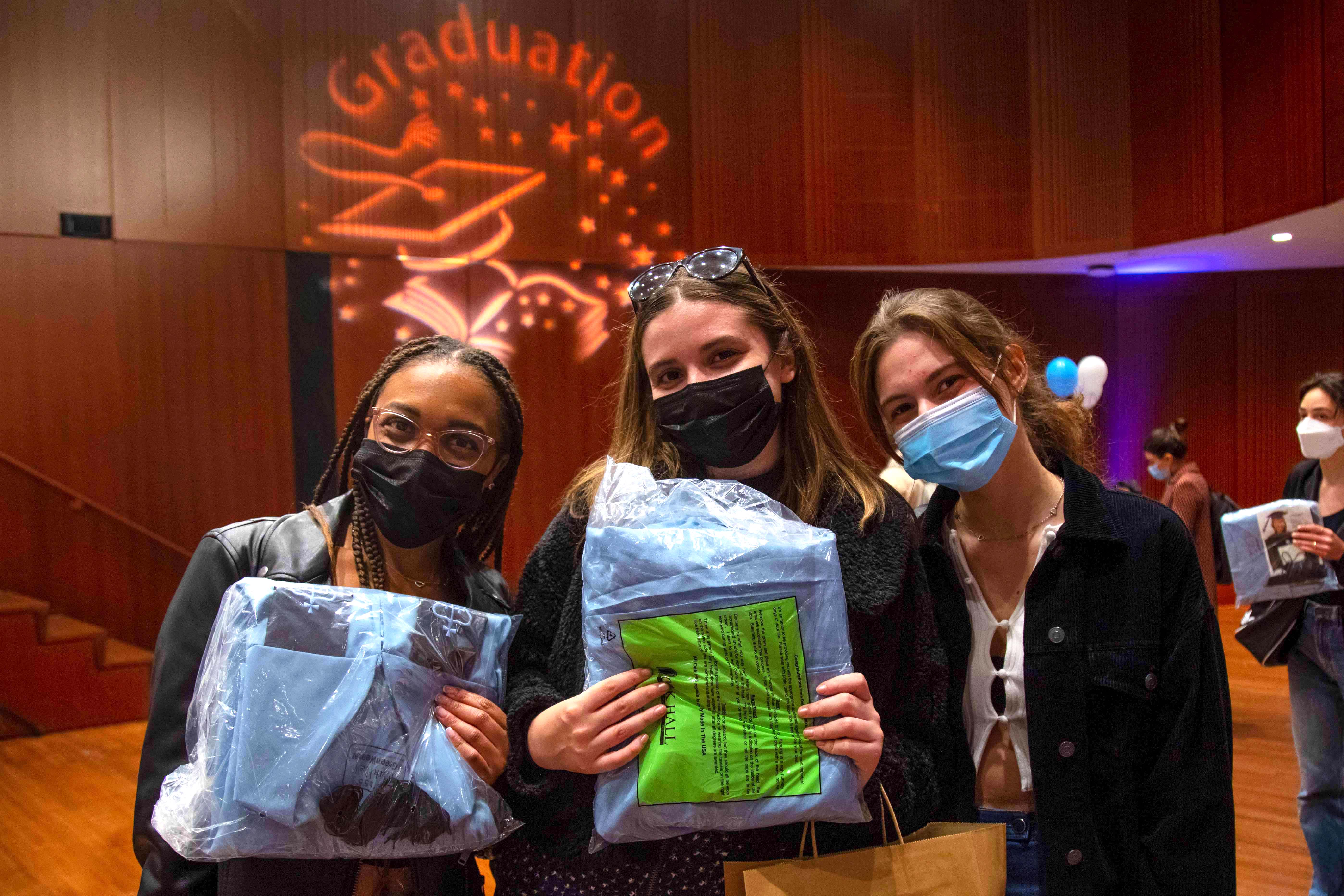 Three students in the Diana Center, two of whom are holding clear bags with their caps and gowns.