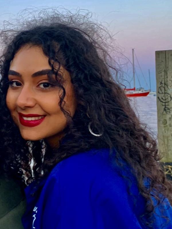 photo of curly haired young woman smiling. she's wearing pinky red lipstick. there is a pink blue sunset in the sky behind her and she is sitting on a wooden dock.