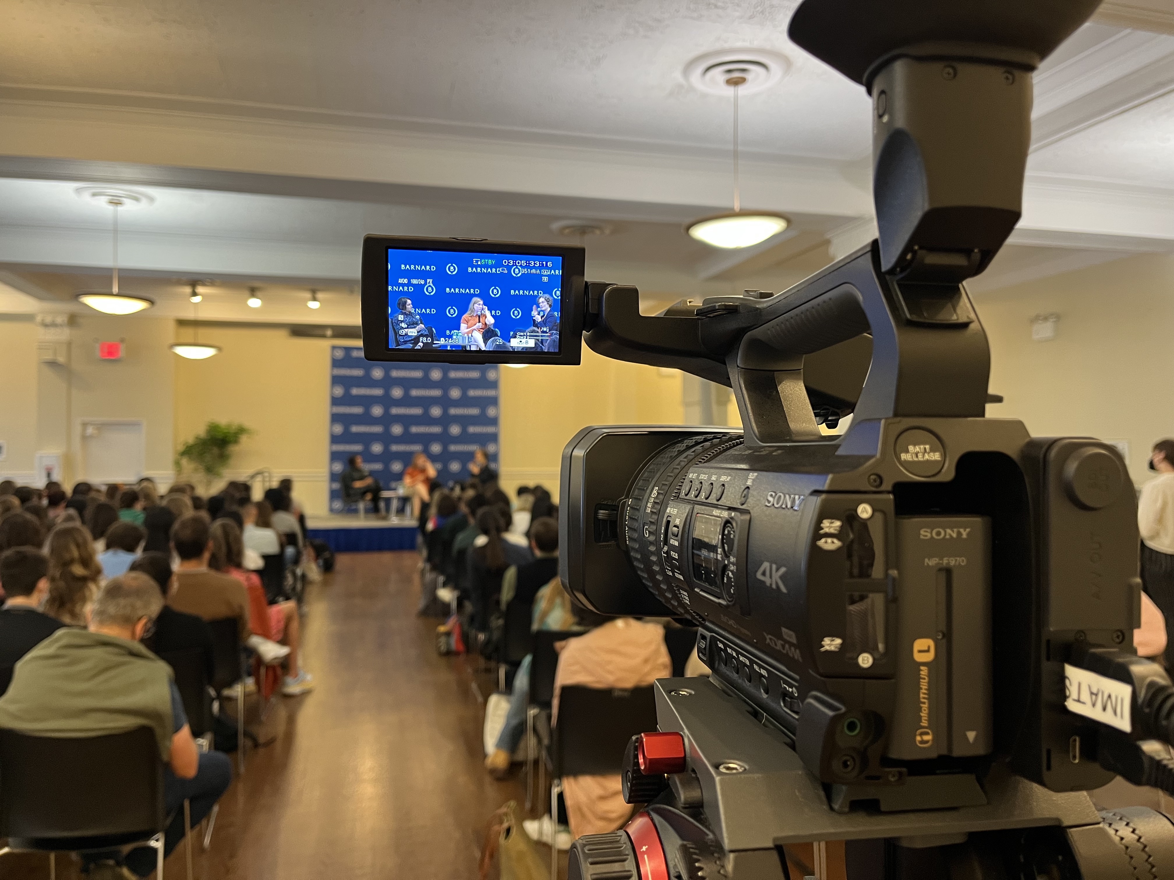 A video camera facing a stage with audience members in a conference room