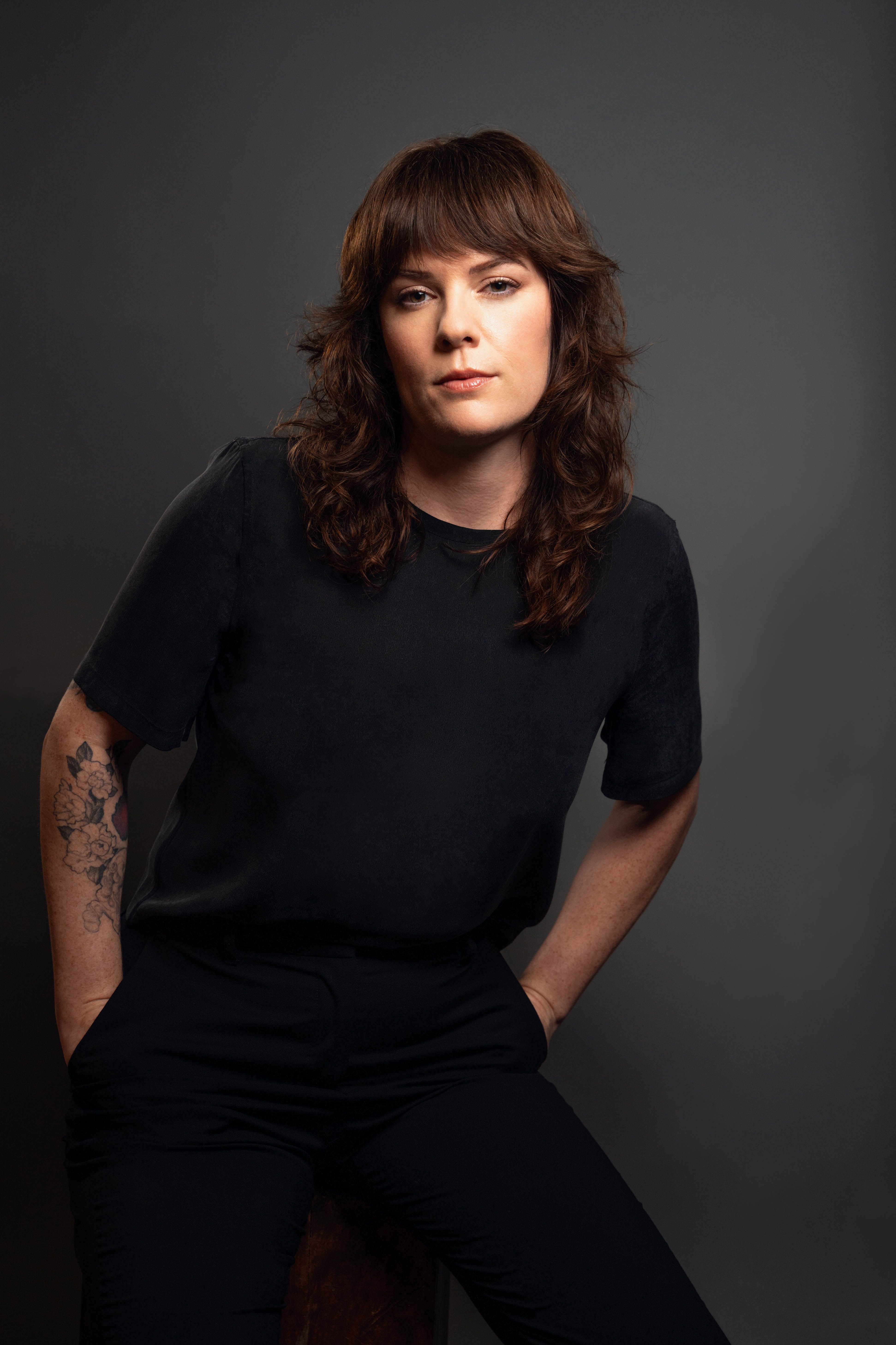 Woman wearing with brown hair and bangs, standing against a dark gray background and wearing black clothing.