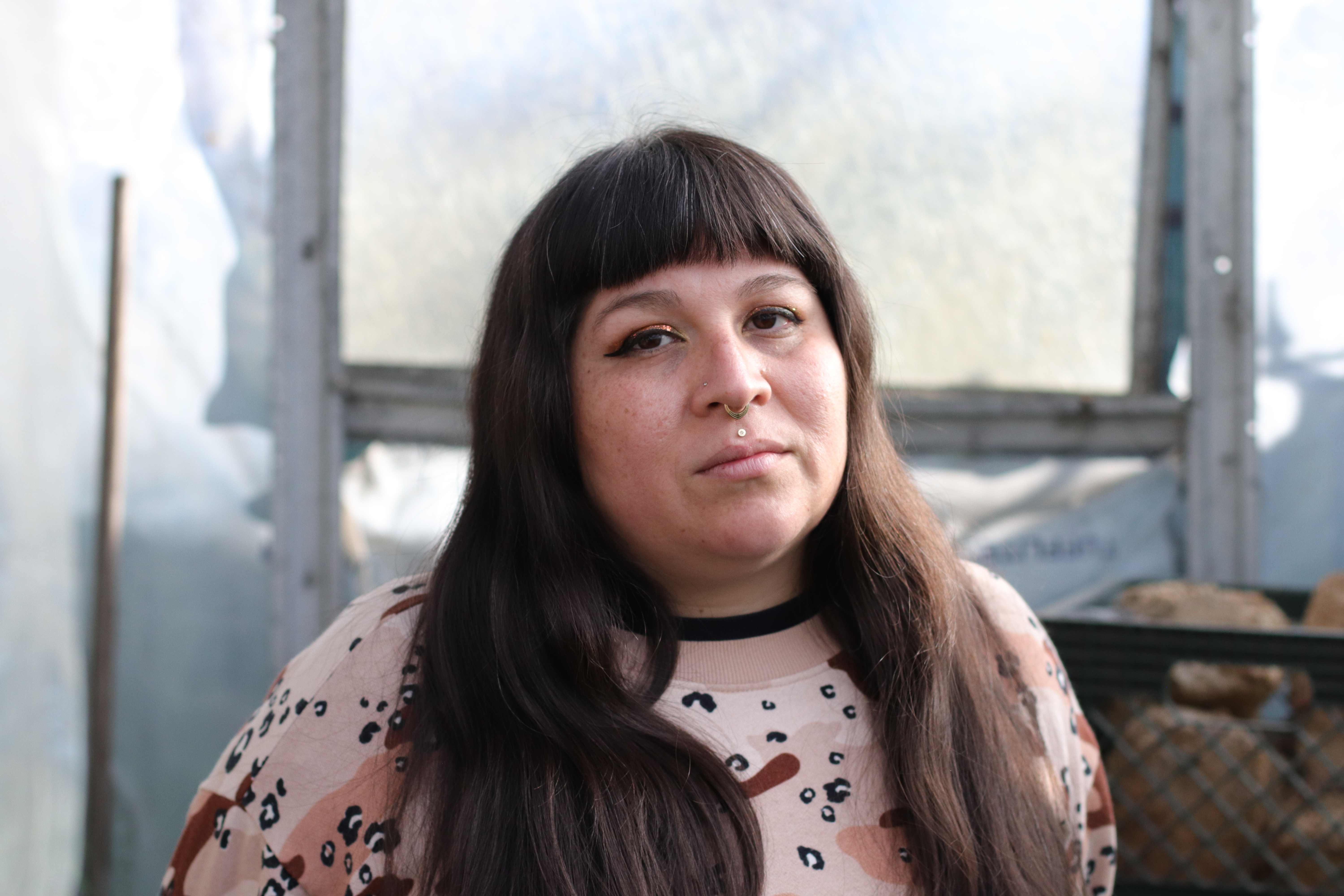 Person with long brown hair, bangs, and a nose piercing, wearing a pink patterned shirt