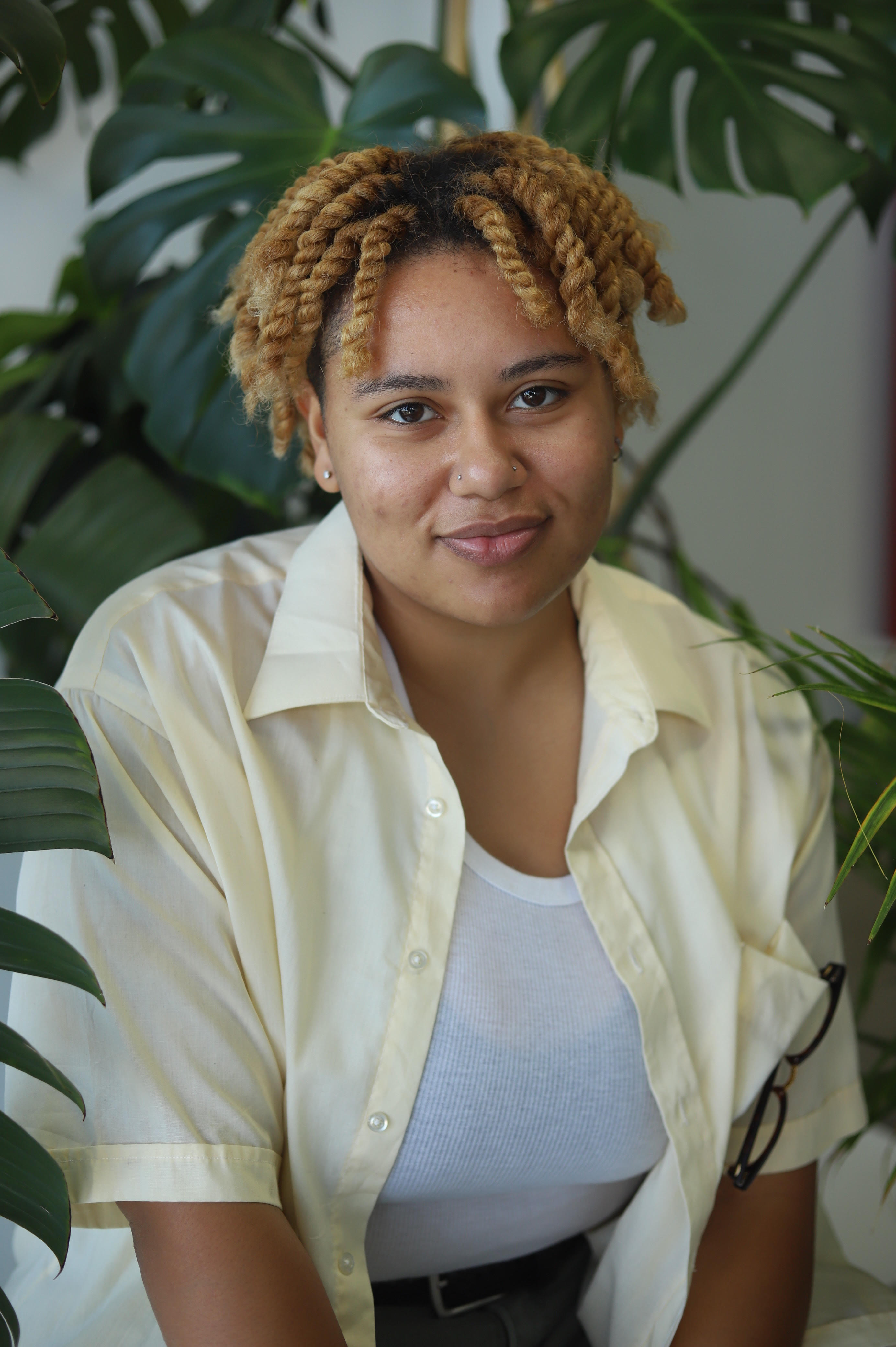 Person with short, light-brown hair worn in twists, wearing nose piercings, and a yellow shirt over a white tank top. Plant is in the background.