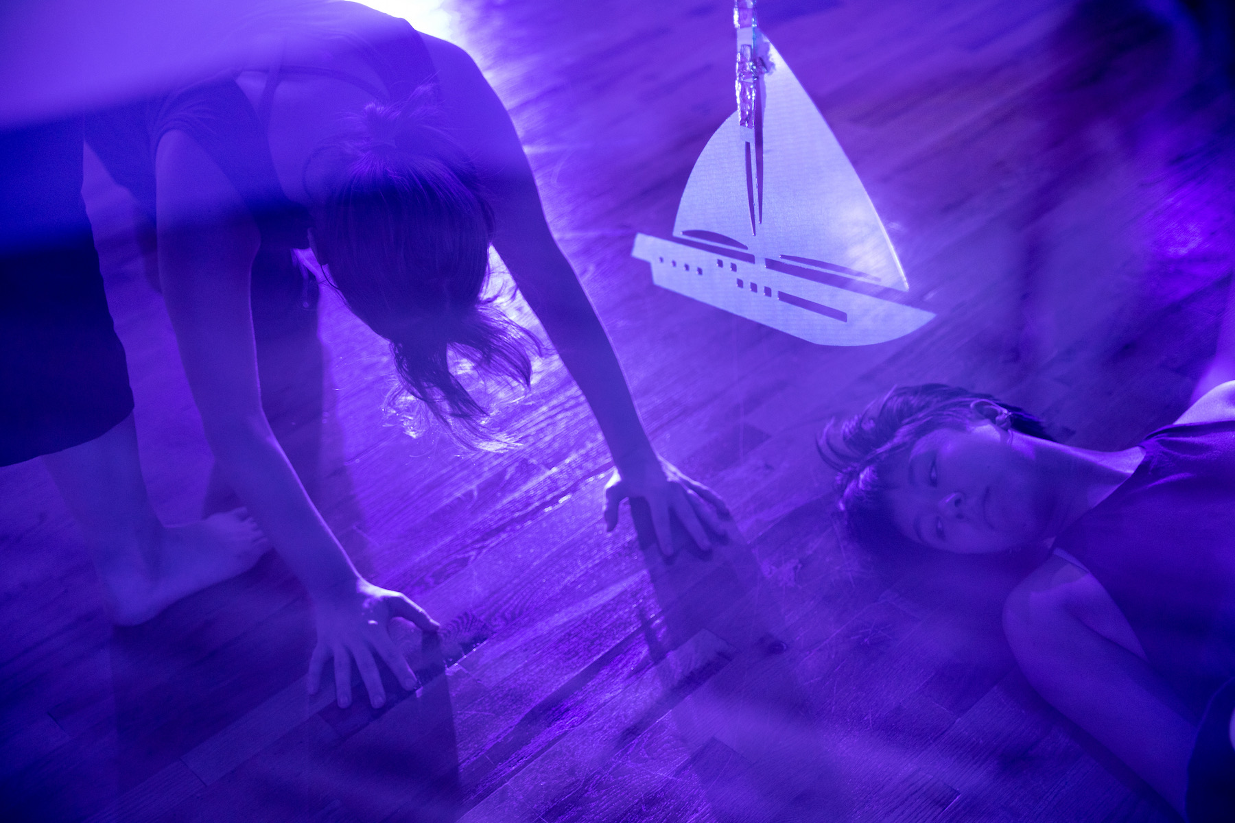 two dancers move through the space in a blue lit room with a metal boat hanging between them 