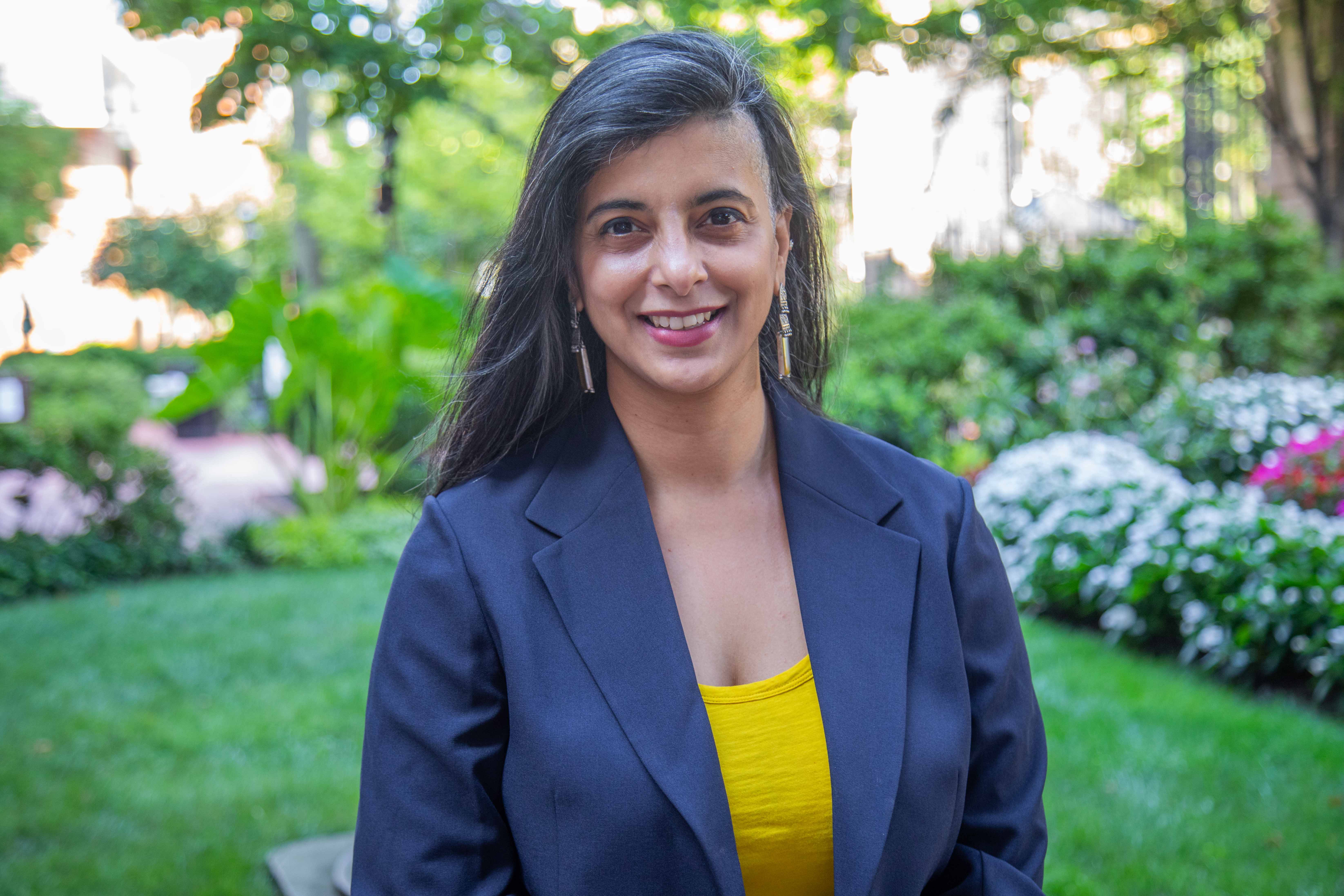 Shayoni Mitra headshot. She is wearing a yellow shirt and black blazer. Campus greenery is behind her