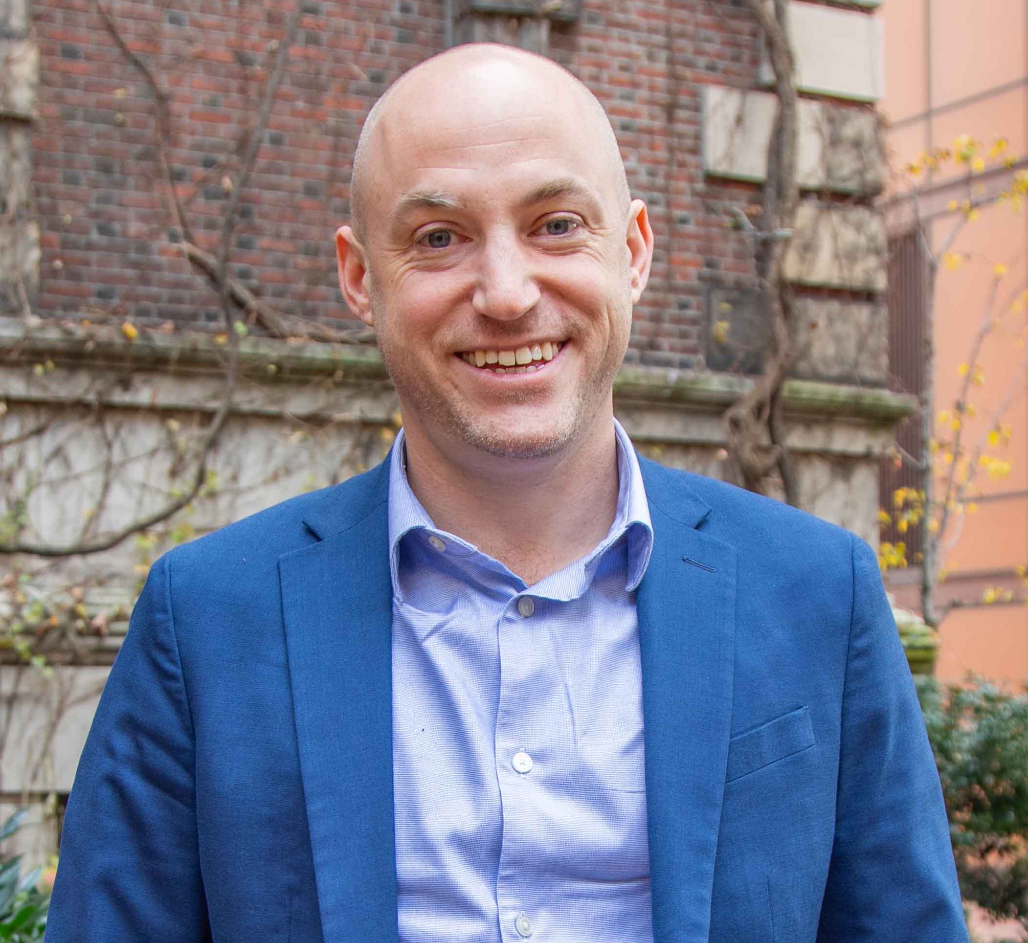 A-J Aronstein smiling in front of a brown building wearing a collared button up shirt (lavender) and a blue blazer