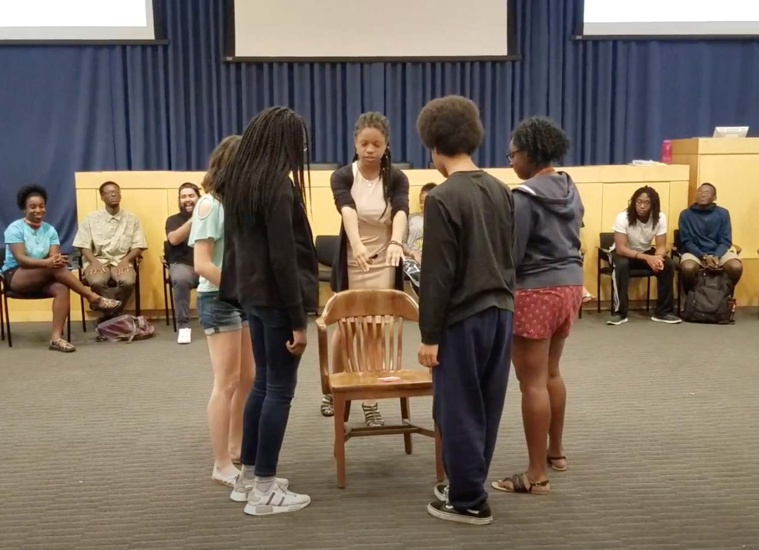 5 students circled around a wooden chair while adults look on