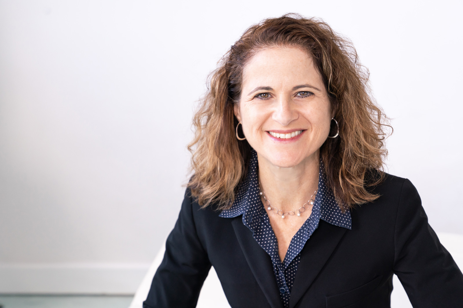 Woman with brown hair wearing black jacket in front of white wall