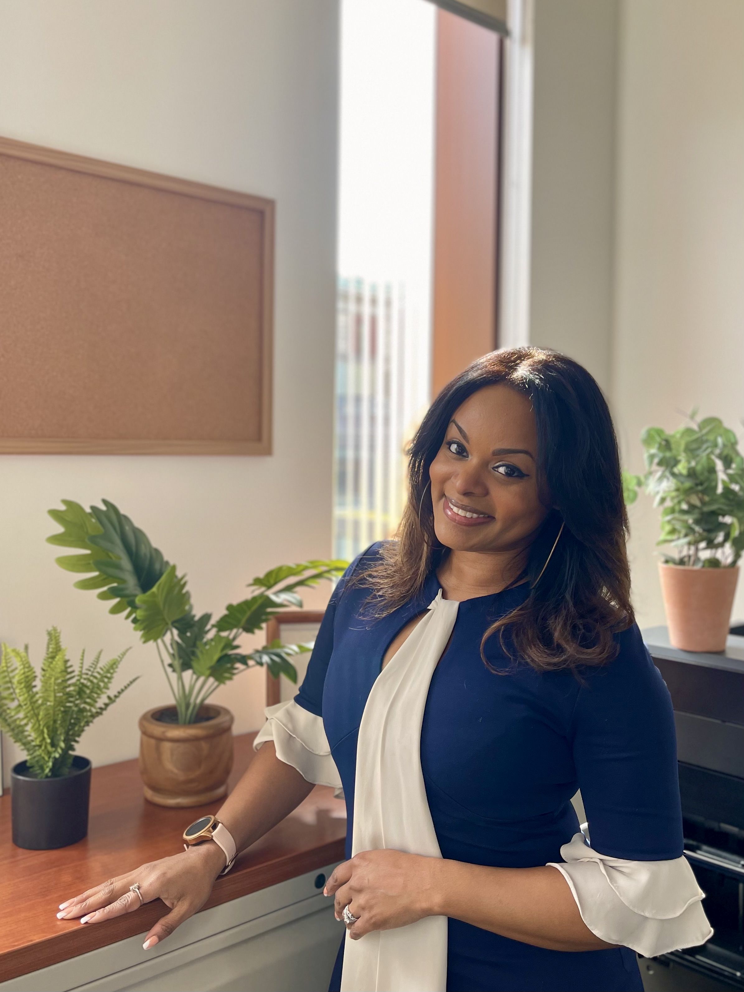 A photo of a woman with long dark hair.  She is standing in front of a sunny window with plants and is smiling. She is wearing a blue dress with off white ruffle accents at the collar and cuffs