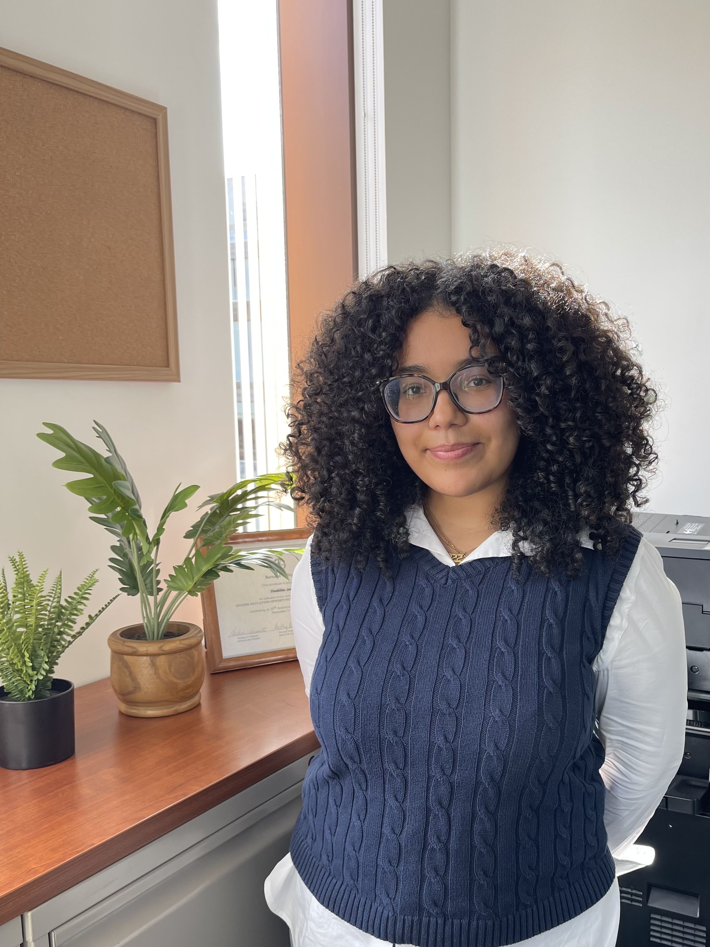 A photo of a woman with curly dark shoulder length hair and glasses stands before a sunny window with plants.  She is wearing a white collared shirt and a dark blue sweater vest. 