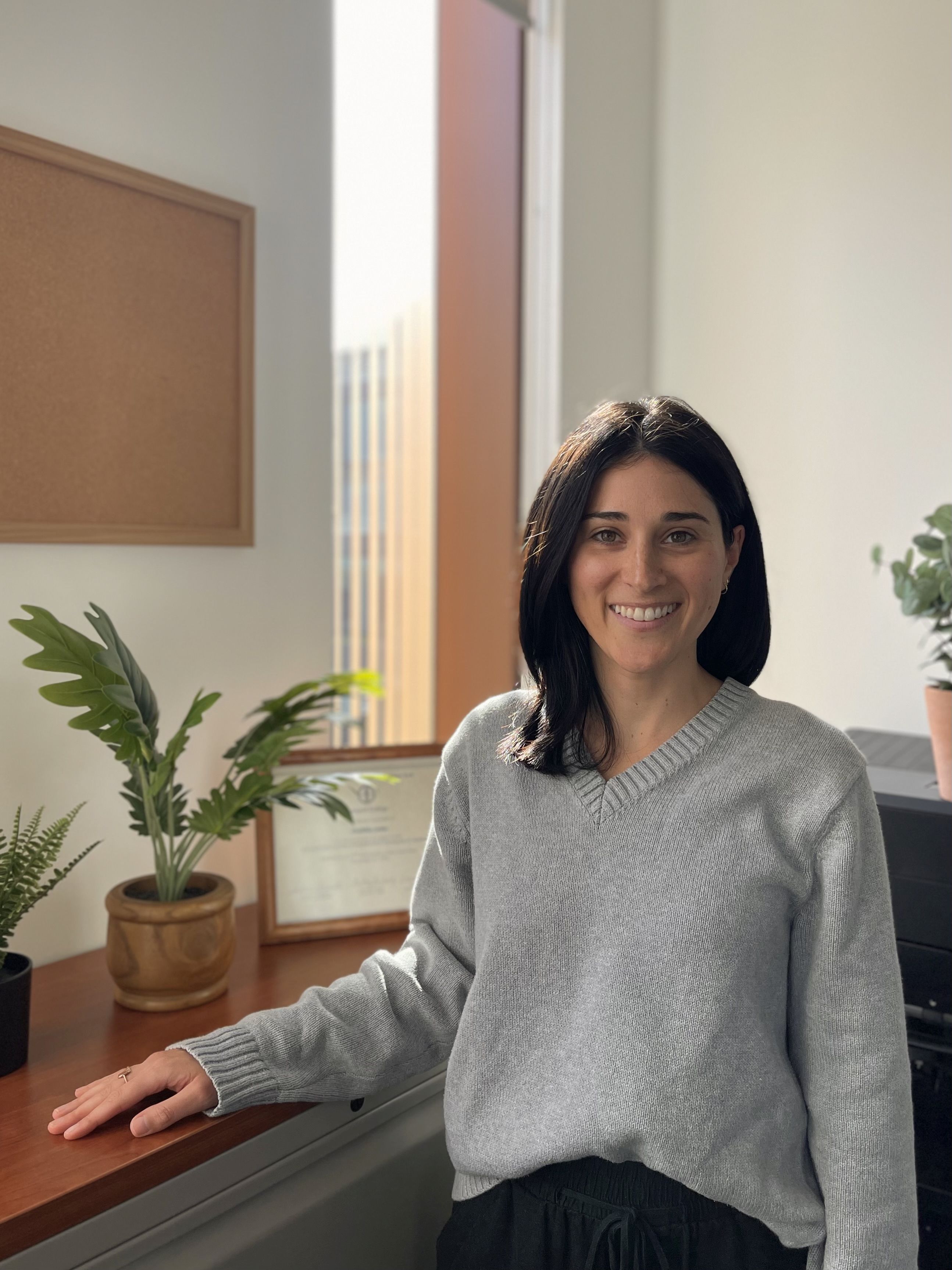 A woman with dark, straight, shoulder length hair stands before a sunny window and plants.  She is wearing a grey turtleneck sweater and is smiling 