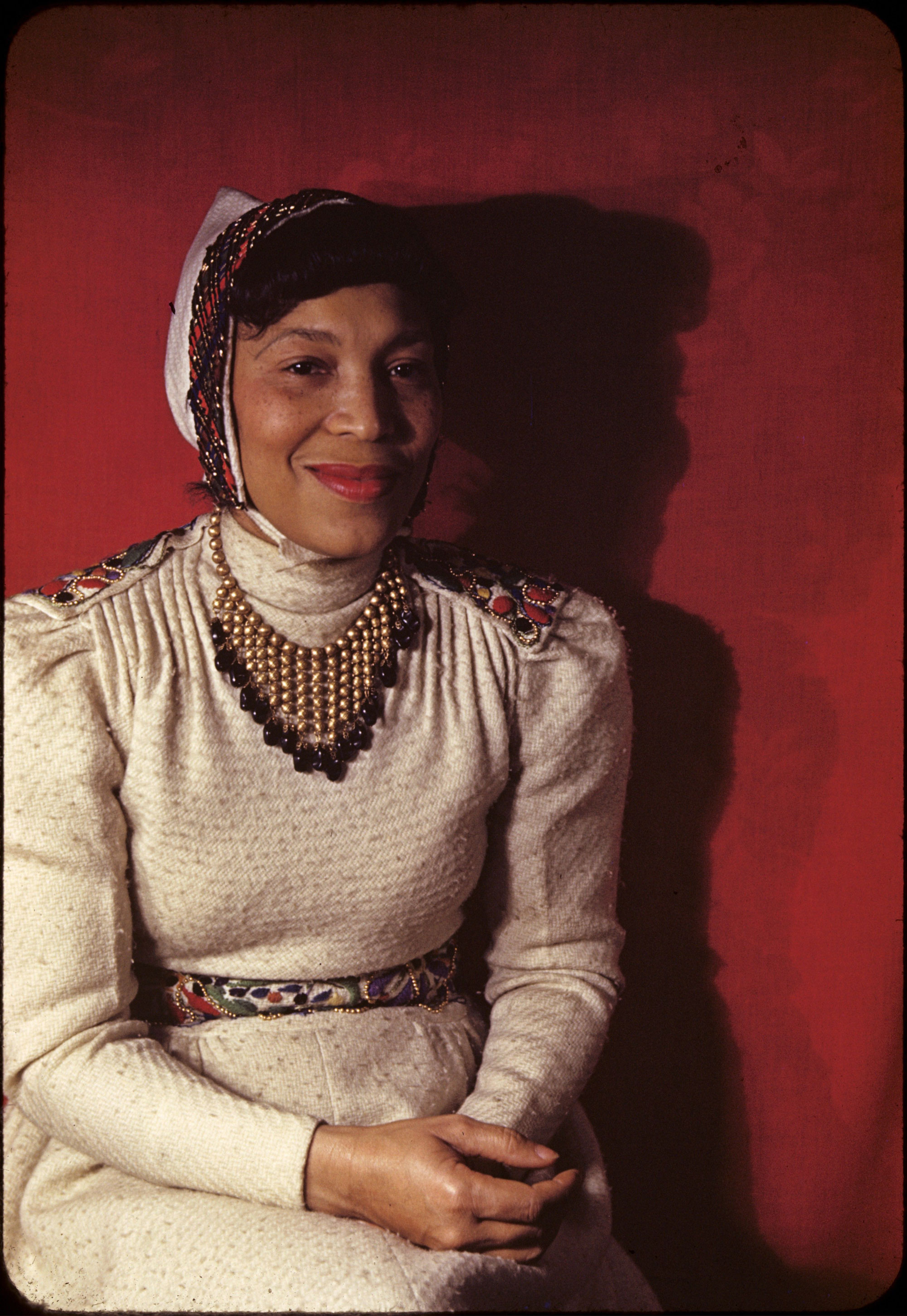 Woman in a sitting pose in front of red wall wearing tan dress and head ornament