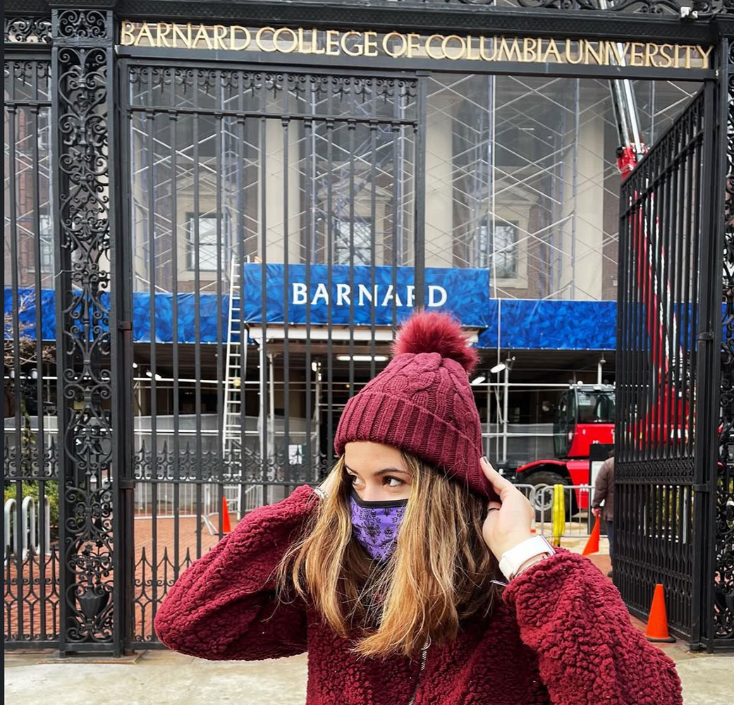 Young woman dressed in maroon colored hat and sweater wearing a mask standing in front of Barnard College