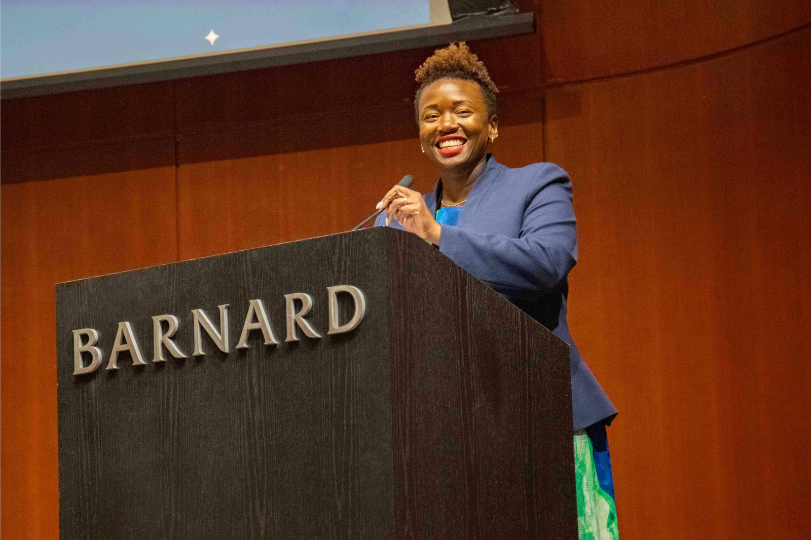 Woman wearing blue jacket stands at podium reading BARNARD
