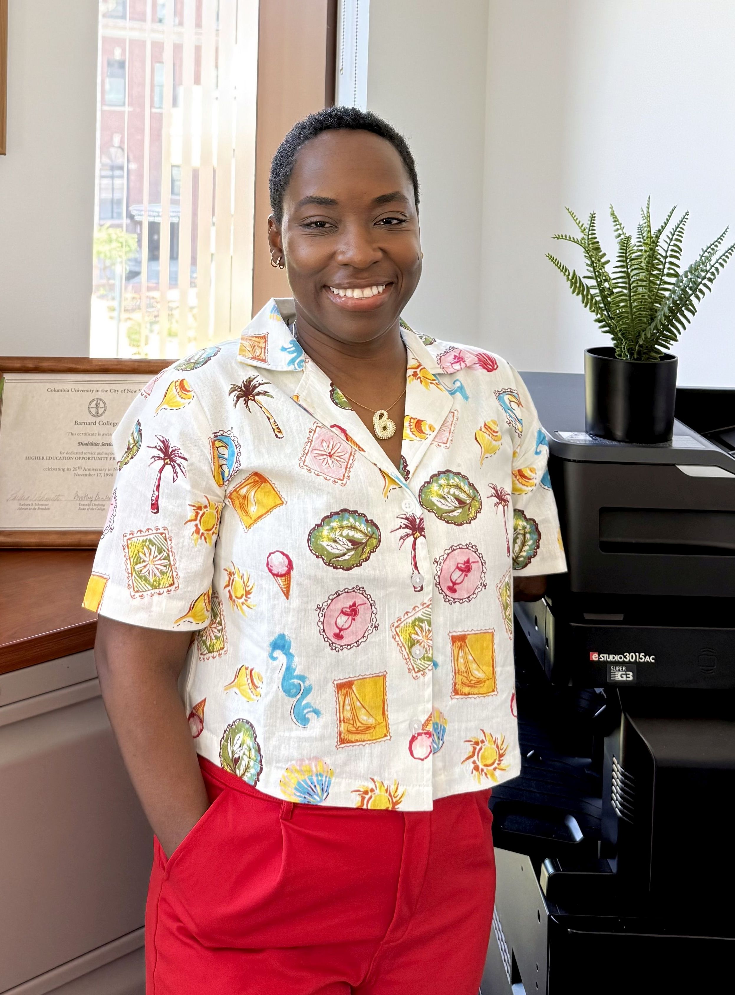 A woman with close cropped dark, curly hair stands before a sunny window and plants. She is wearing a white short sleeved shirt with a colorful pattern and red pants. She is smiling.