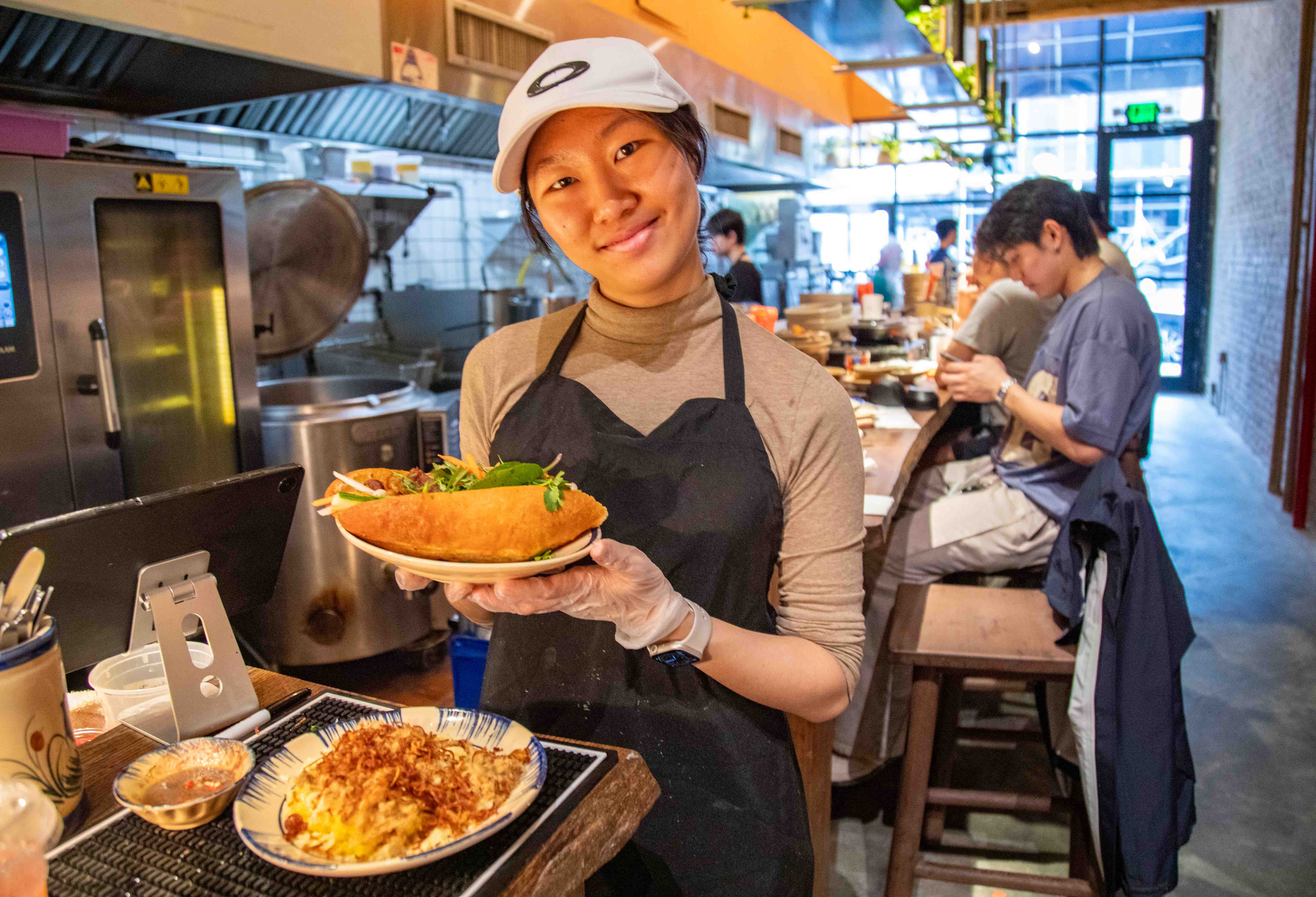 Young woman wearing a cap and apron inside of a kitchen holding a bahn mi