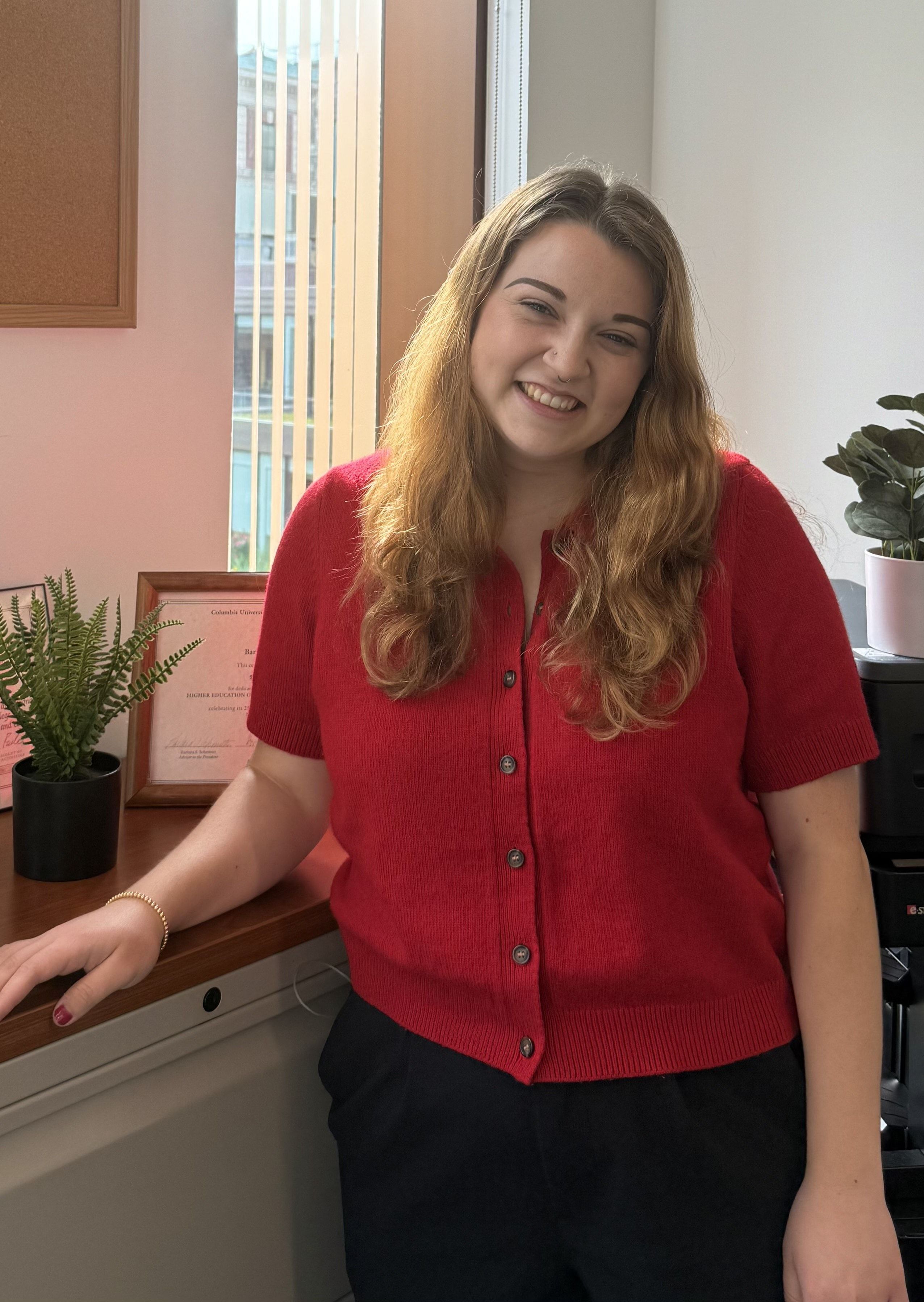 A woman with straight reddish brown hair stands in front of a sunny window with her hand on a cabinet.  She is wearing a bright red short sleeved sweater and black pants and is smiling at the camera.