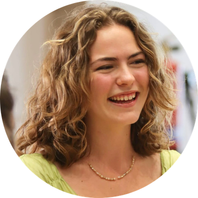 Elsa wears a lime green shirt and has curly hair. She looks to the right and smiles with her mouth open.