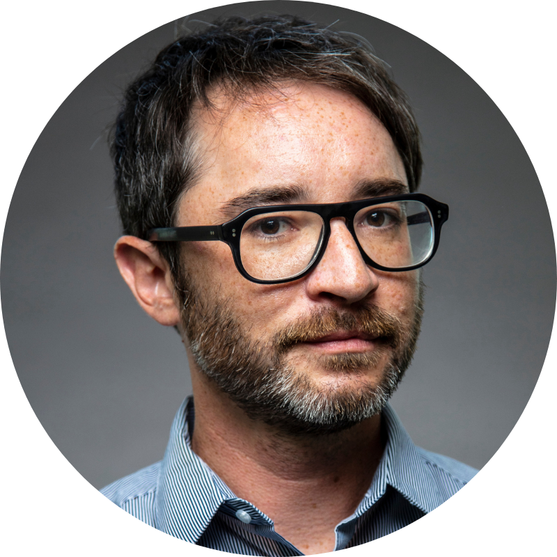 Nathan Gorelick wears dark rimmed glasses and a blue collared shirt. He has a beard and short hair. 