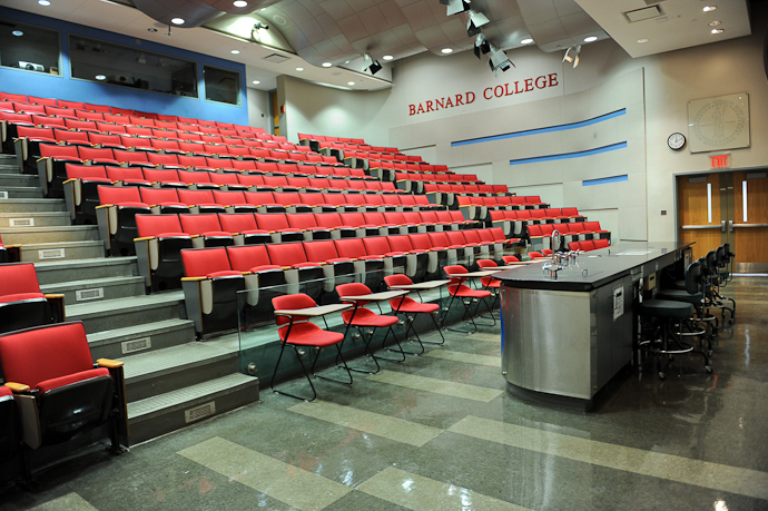 Shows a large auditorium with tiered stadium seating with red chairs and a long laboratory table with stools at the front