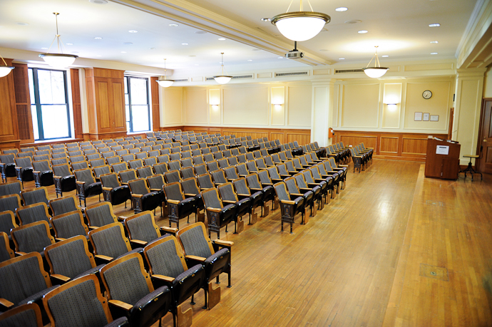 Shows a large lecture hall with wooden floor and walls, and built in padded seats. There is a small wooden stage at the front with a podium.