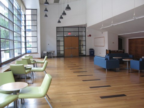 A picture of the Altschul atrium showing wood floors, a wall of glass windows, and doors at the center of each wall. The room has couches and tables.