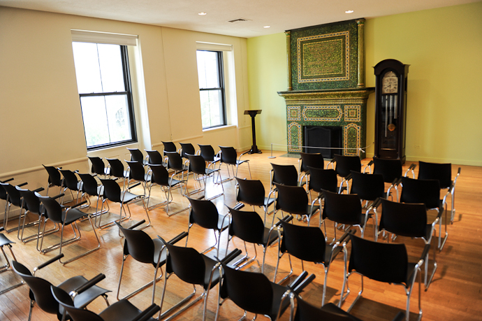 Shows a small room with wooden floors set with black chairs facing a large Tiffany fireplace.