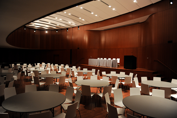 shows oval-shaped room with high ceilings near the stage and warm wooden paneling. The room is set with round tables and chairs, and a podium and panel on a small rectangular stage.