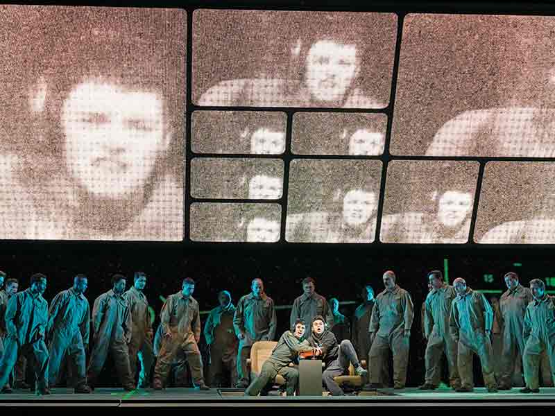 The Met Opera production of Grounded, showing the singers with large digital closeups on screens behind them