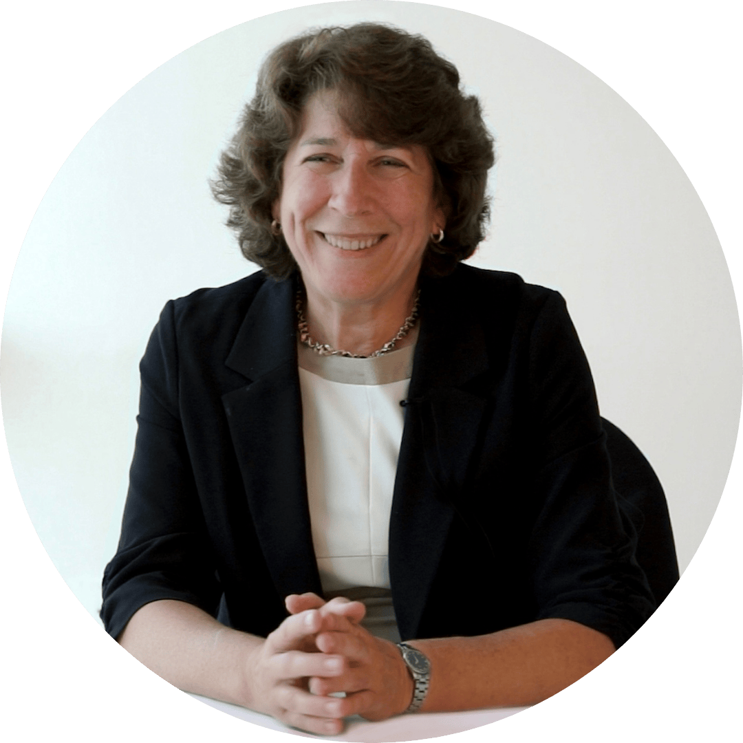 Professor Kathleen McKeown smiles with her hands folded on a table. She has curly hair, and wears a white shirt and black blazer.