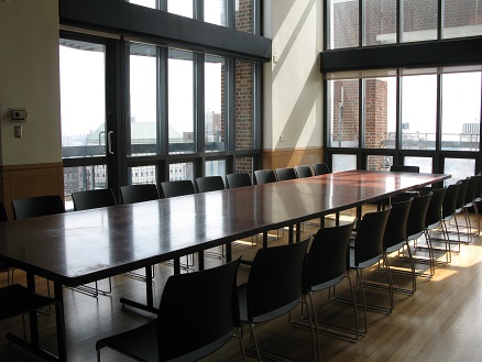 shows room with tall ceilings and extensive floor-to-ceiling windows, set with a wooden rectangular table and black chairs.