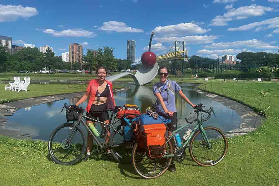 Brenna and her biking companion pose with their bikes in front of a pond with blue sky and fluffy clouds behind them