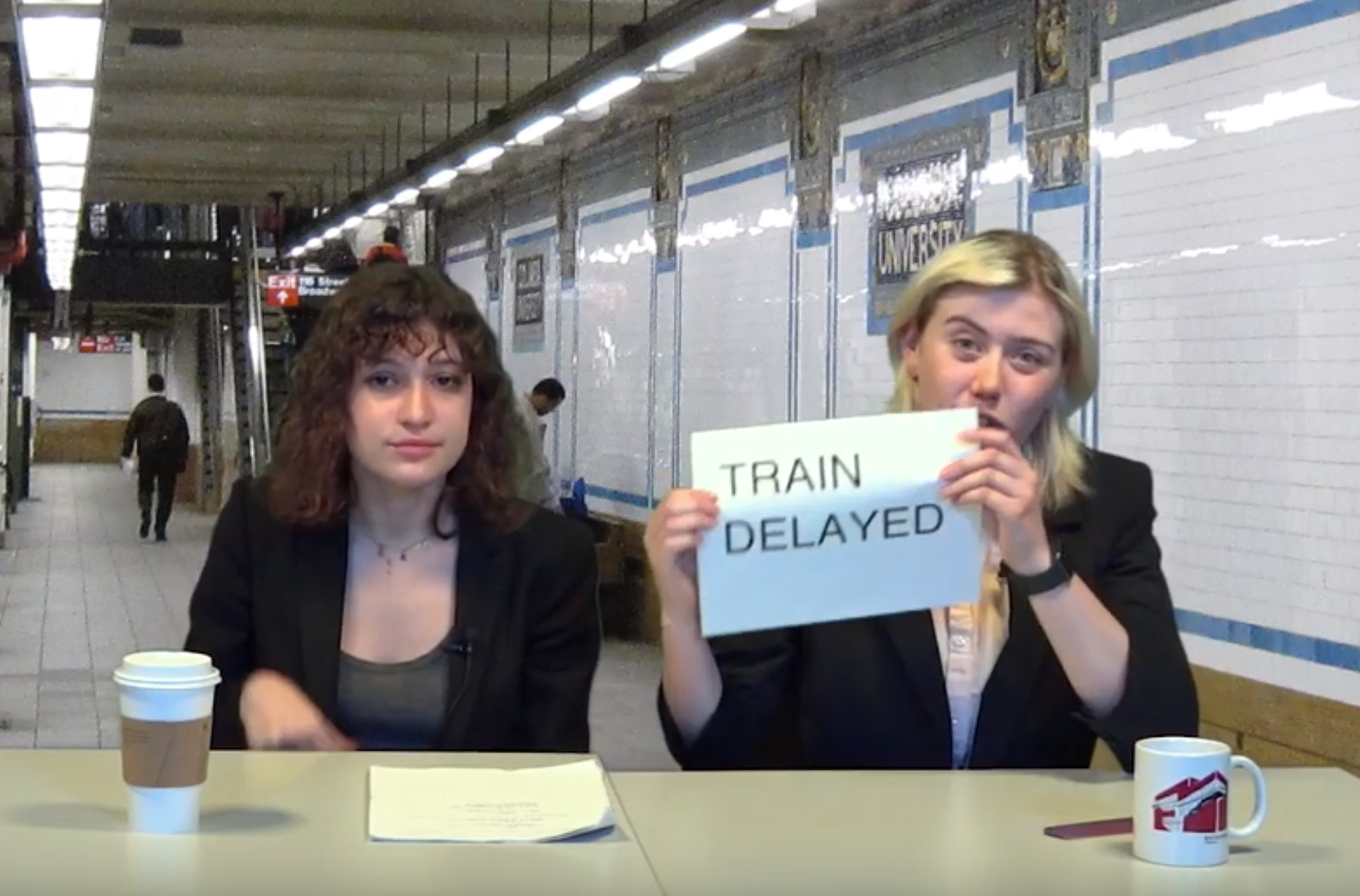 Students sitting at desk at Columbia subway station; one is holding a sign that says 'train delayed'