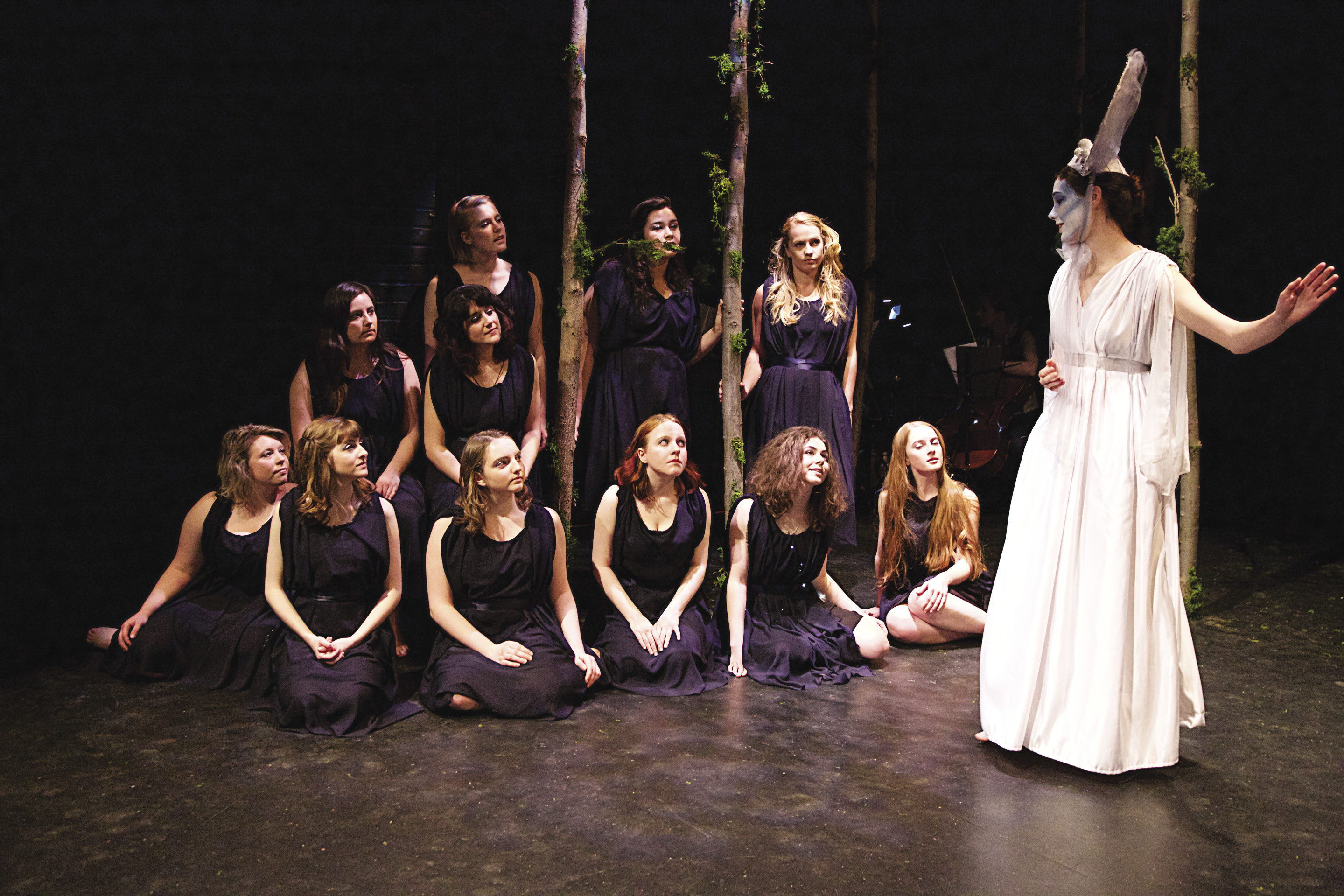 A woman in white walks past a group of women in black, kneeling on a stage
