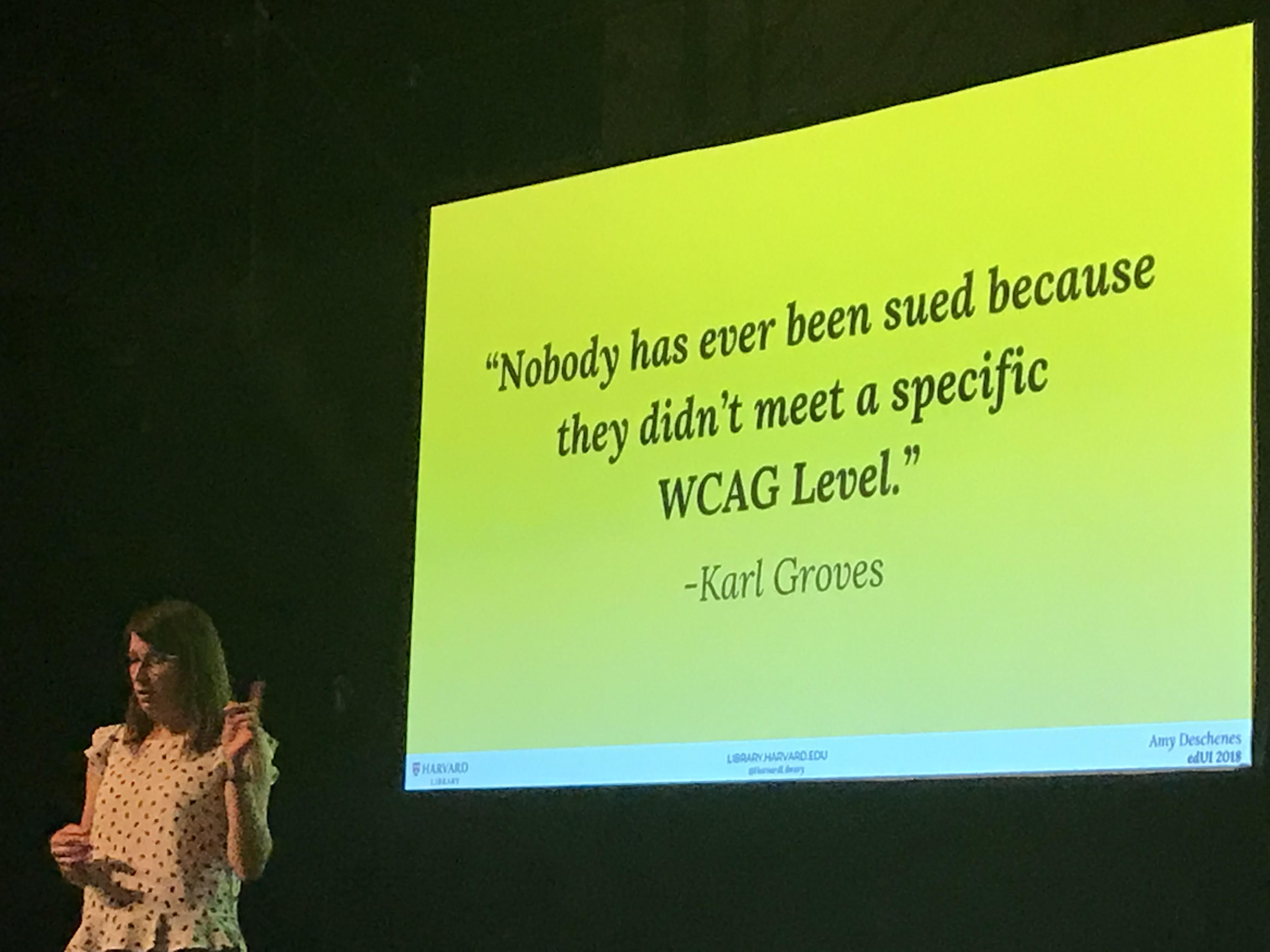 A female lecturer in front of a slide that has the quotation '"Nobody has ever been sued because they didn't meet a specific WCAG Level." -Karl Groves' written on it. 