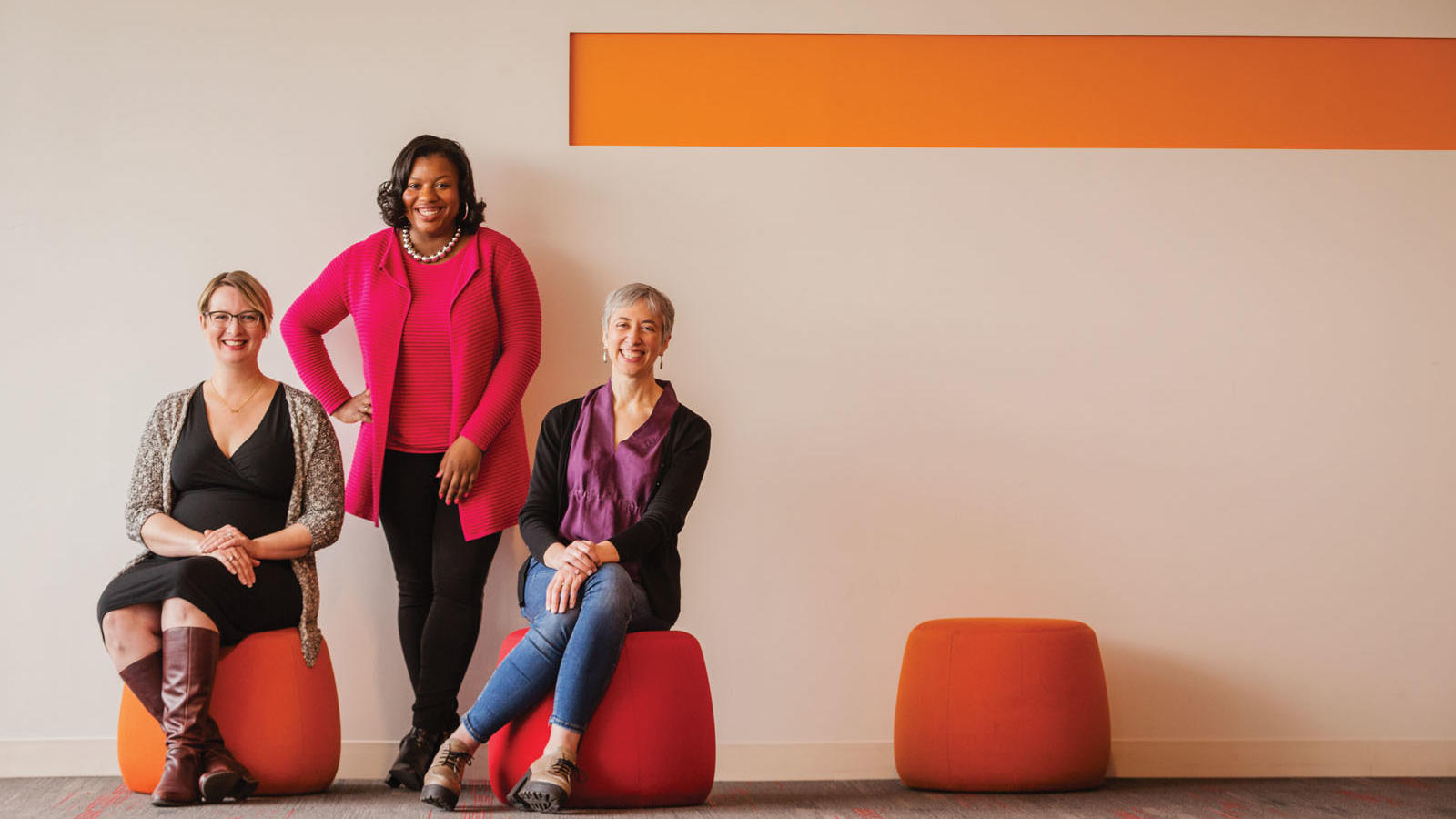 3 women facing forward, two sitting on orange stools