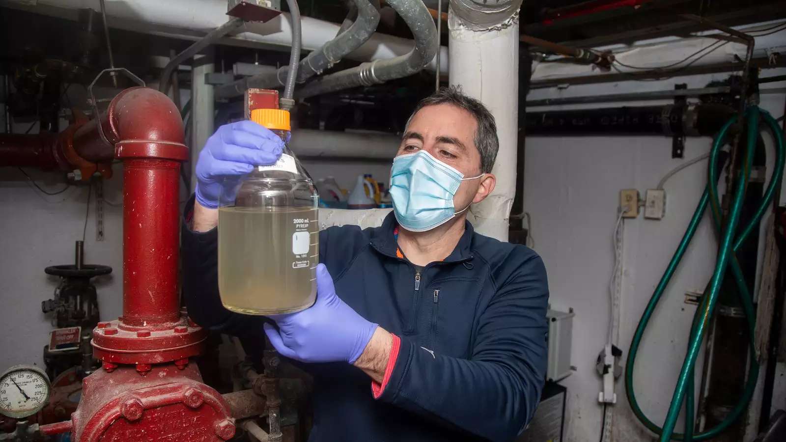 Man in mask and surgical gloves holding jug of wastewater sample in building basement