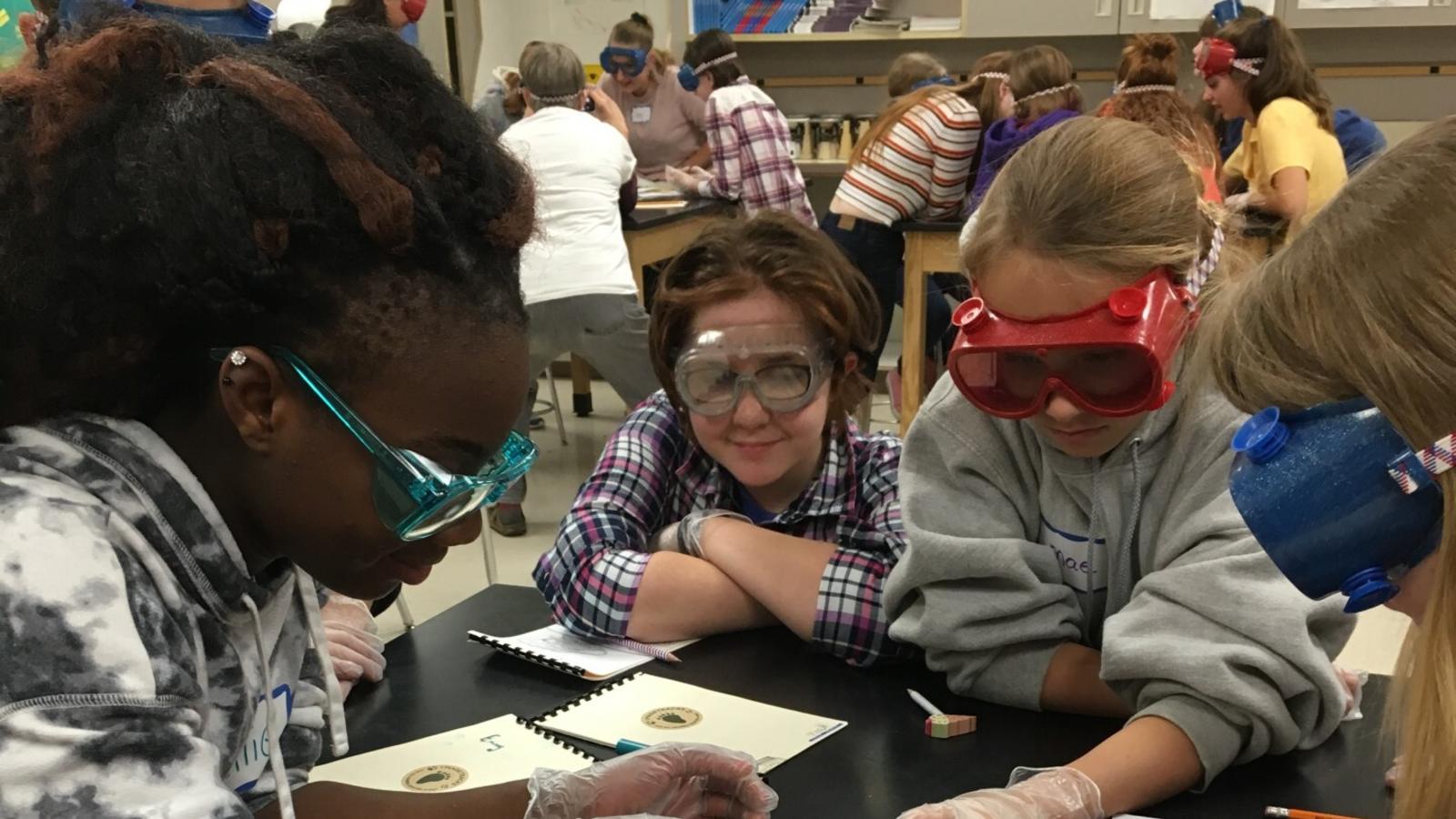 Six young girls crowd around a science lab table working on an activity. Other tables of children can be seen working in the background.