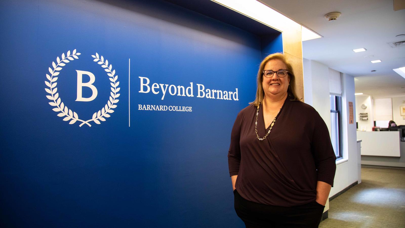 Christine Valenza Shin stands in front of a blue wall with the Beyond Barnard logo on it, located in the Beyond Barnard office