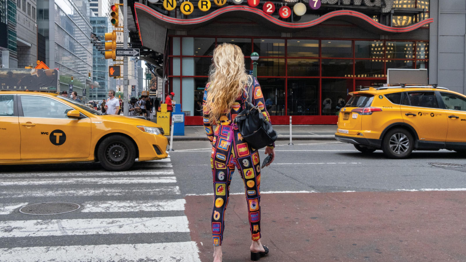 Woman crossing a street in Times Square; her colorful jumpsuit matches the subway sign