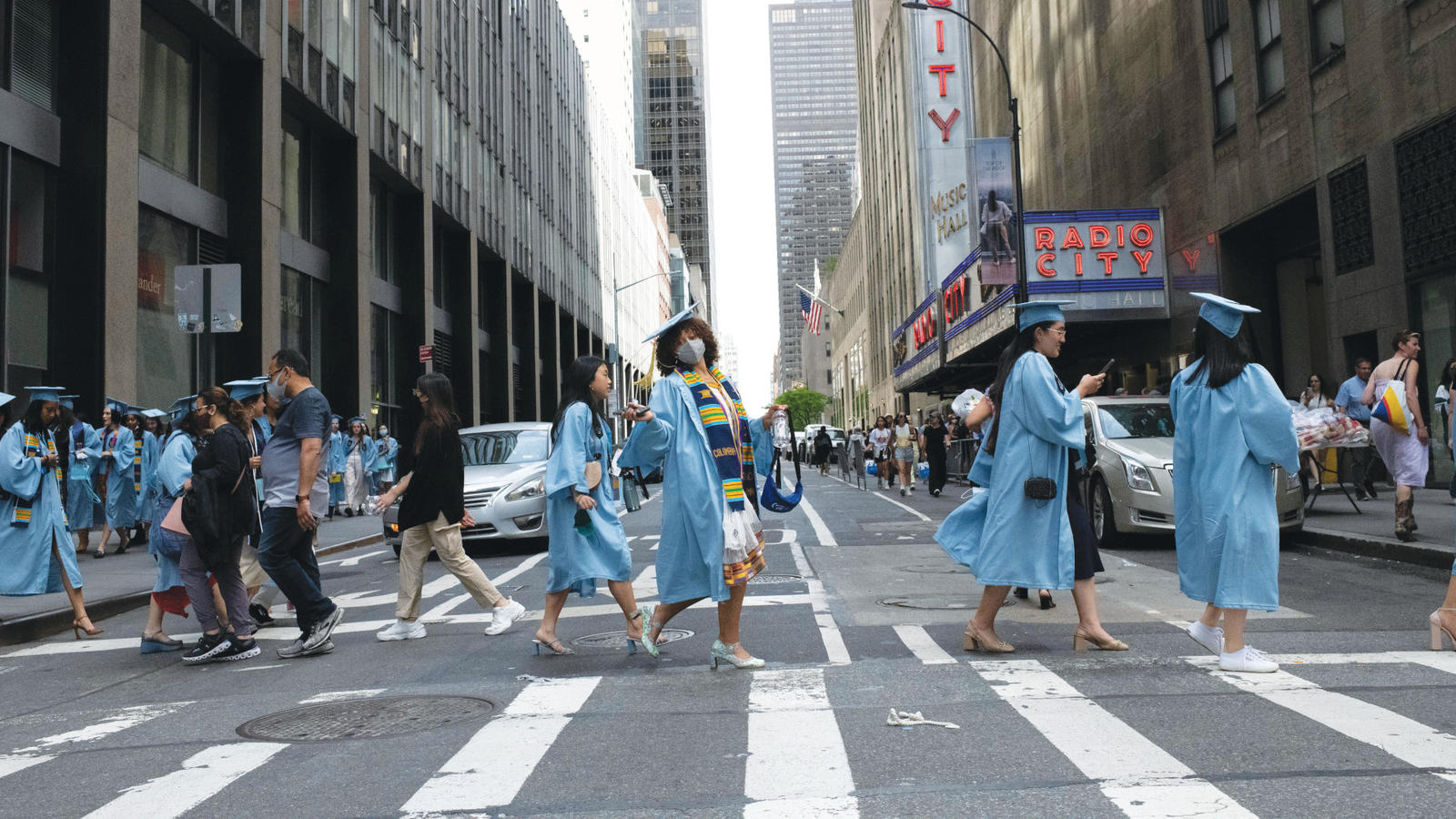 Barnard women in graduation gowns stride across a city crosswalk