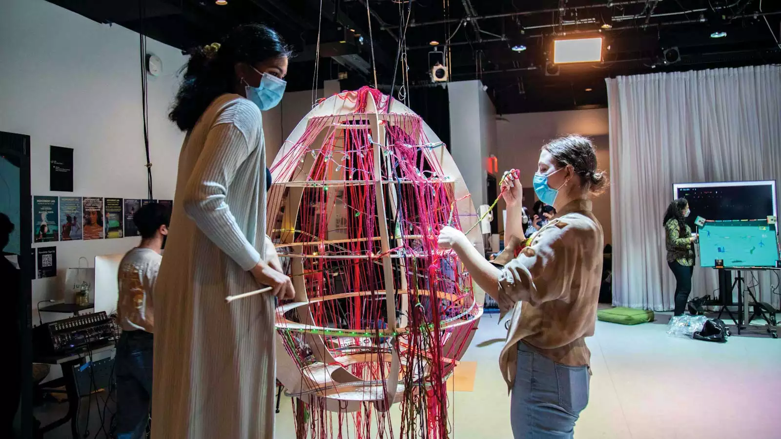 2 students work on a large, egg-shaped object weaving colored yarn