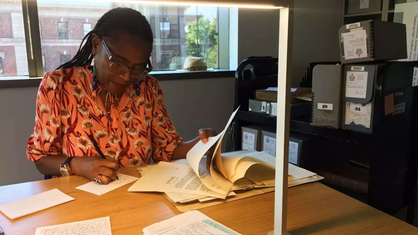Researcher seated desk and taking notes on index card while holding archival materials.